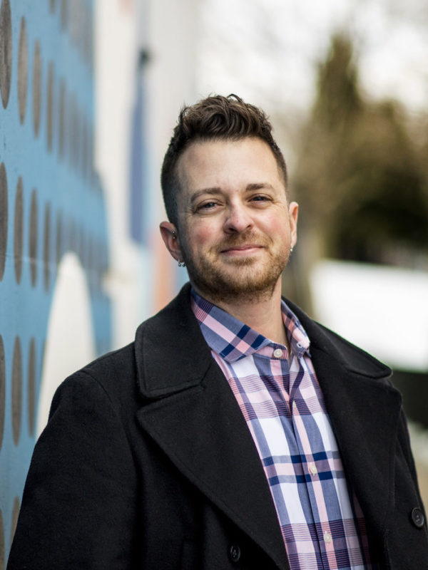 Headshot of a white-presenting individual with short hair and facial hair, wearing a pink and blue plaid shirt under a winter coat. They are smiling into the camera