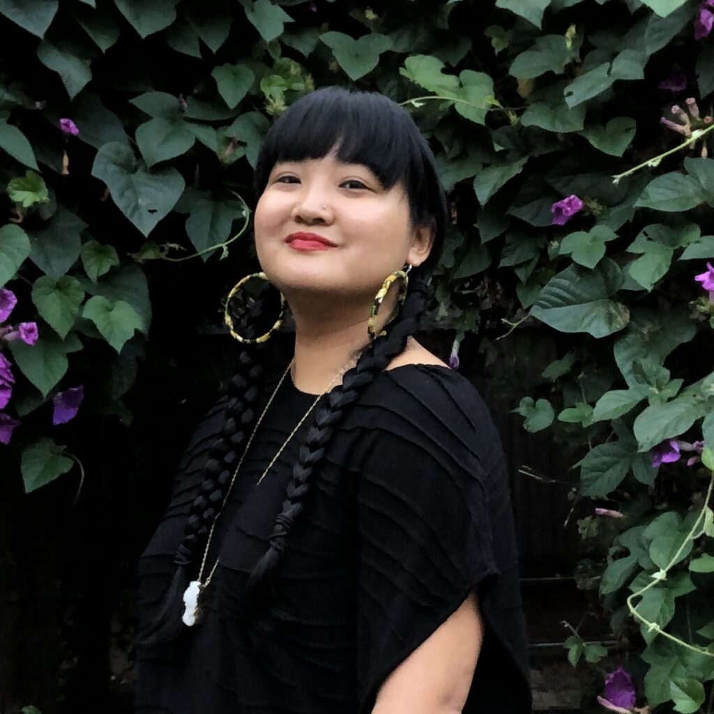 Female presenting Asian woman with two breads and large hoop earrings wearing a black shirt. They are standing sideways and smiling into the camera in front of greenery