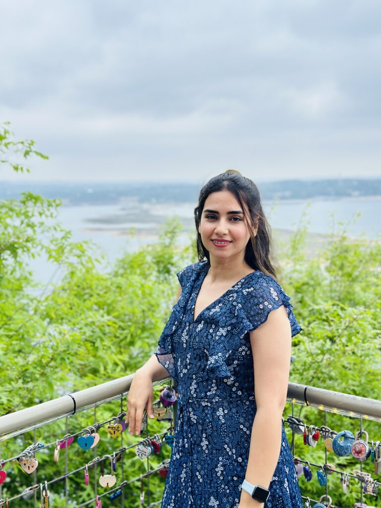 A female-presenting individual with dark hair leans on a railing, smiling into the camera. Green leaves and water are visible in the background
