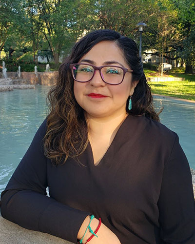 A female-presenting person wearing glasses looks into the camera, their arm resting on a wall. Water is visible behind them